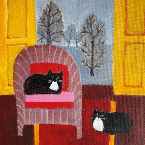 Two black and white cats, one sitting on a chair and one on the carpet in front of an open door with trees outside.