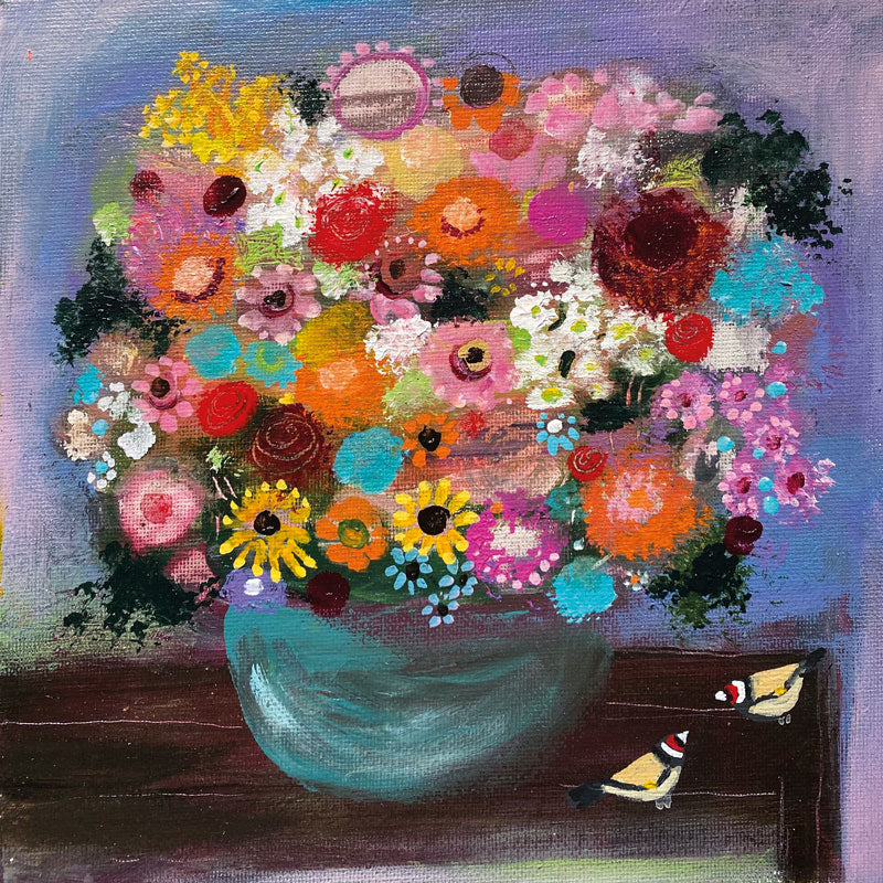A vase full of colourful flowers with two Goldfinches on the table next to it.