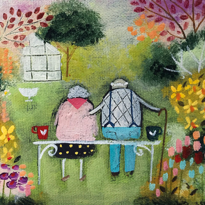 An old couple sitting on a bench in a garden  with cups of tea. He has his arm around her.